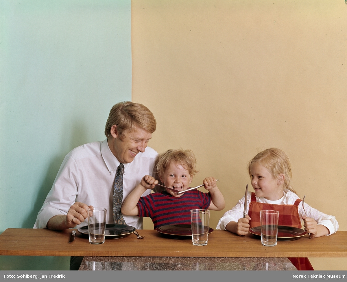 En mann og to barn, en gutt og en jente, sitter ved et bord.Foran seg har de tomme tallerkner og glass. Det ser ut som en liten familie som venter på middag.