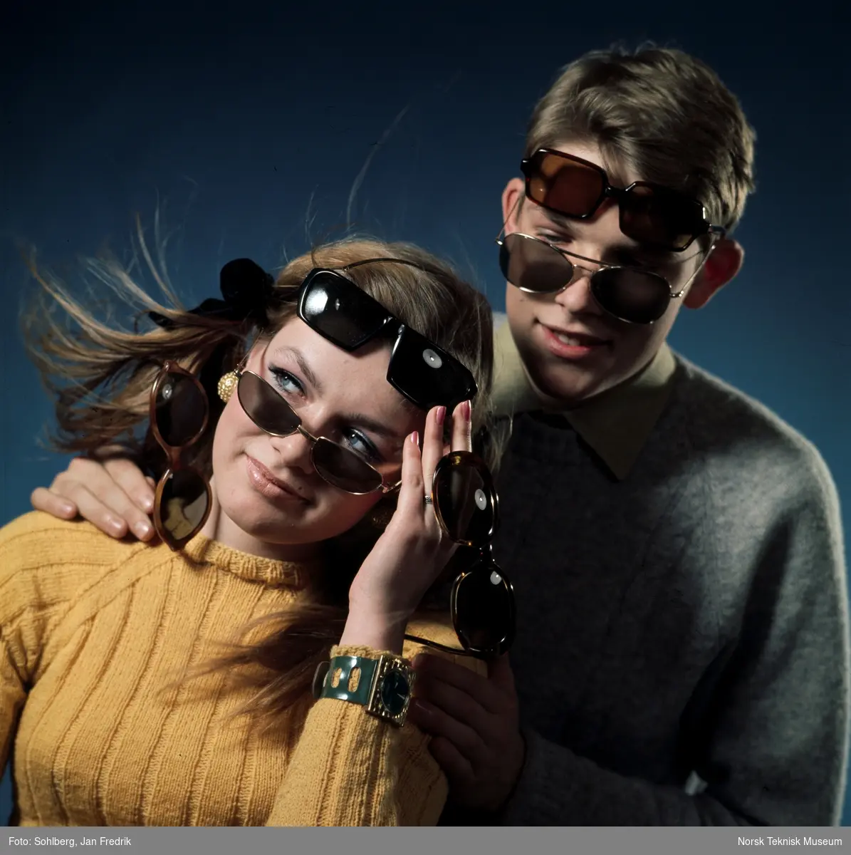 To modeller, en kvinne og en mann, viser fram tidens solbrillemote. Begge har mange par solbriller i forskjellige former på seg. Humoristisk reklamebilde.