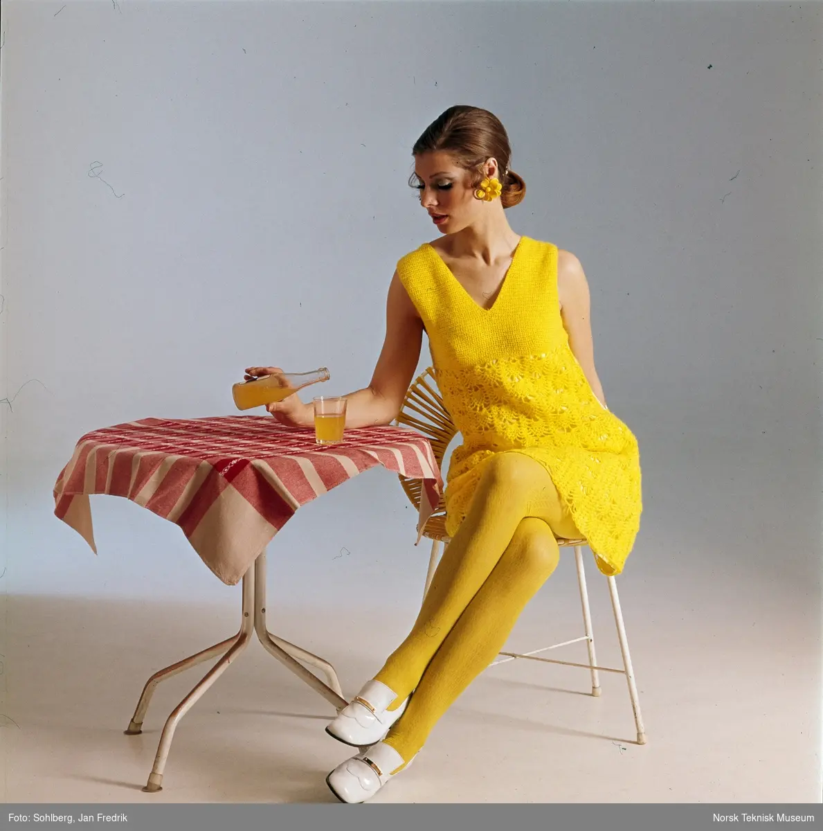 En kvinne i gul kjole og gule strømpebukser, sitter ved et kafebord. På bordet står det en flaske og et glass med gul brus.