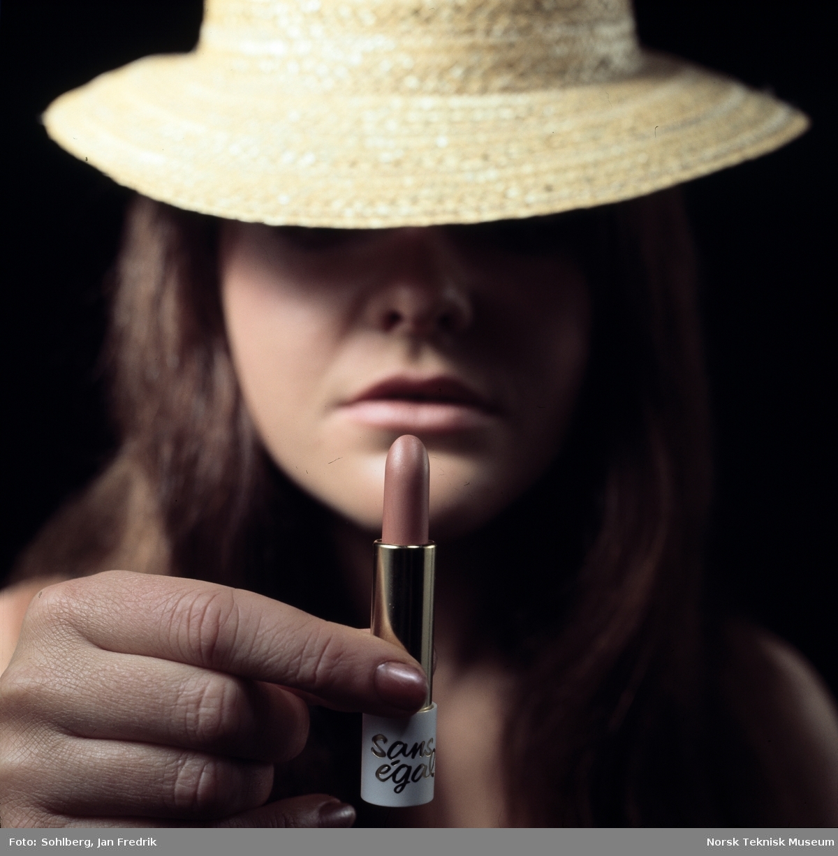 Reklamefoto for leppestift. Nærbilde av kvinne med hatt som holder en leppestift opp foran ansiktet sitt.