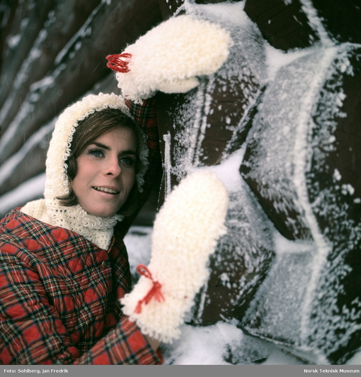 Kvinnelig modell viser vintermote. Hun har en hvit heklet genser med hette, laget av tykt, loddent garn under en skotsk-rutete vattert jakke. Store votter av samme type garn som hettegenseren. Hun lener seg mot en vegg av laftet tømmer.