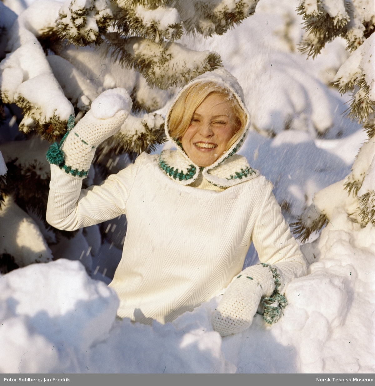 Kvinnelig modell viser vintermote. Hun har hvit genser, hvit strikket hette med grønt mønster og hvite votter med grønne snorer.