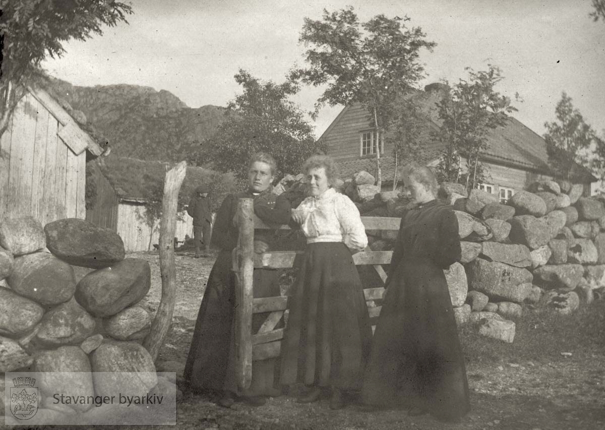 Tre kvinner foran grind og steingjerde. Gårdsbruk i bakgrunnen.