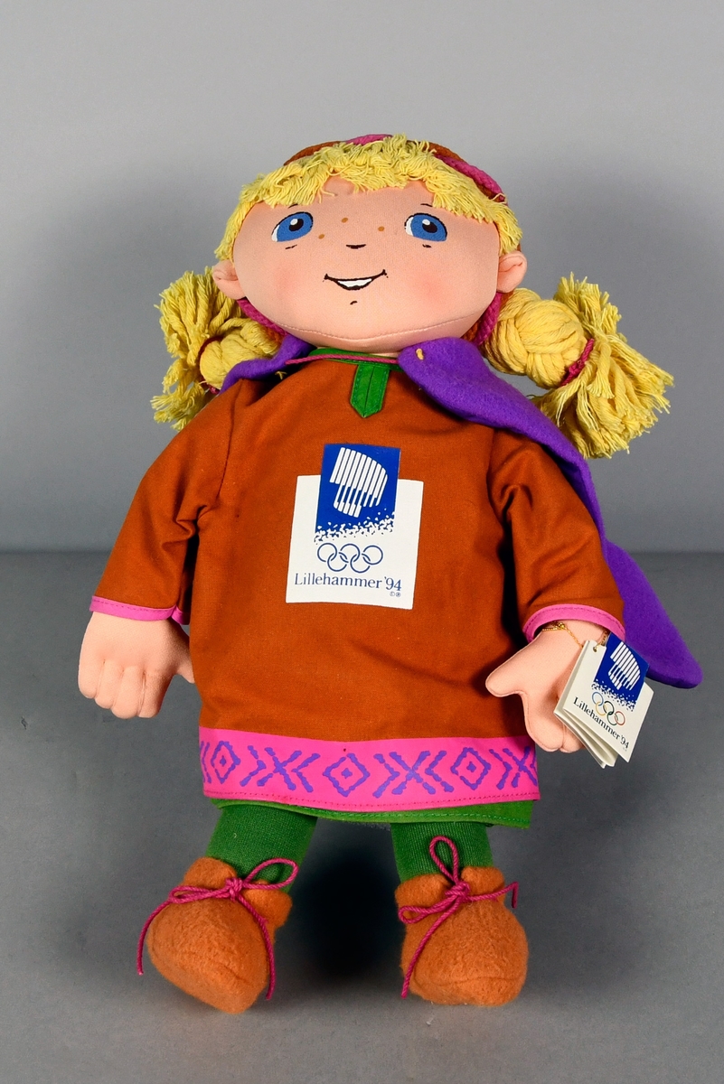 Tøydukke som forestiller maskoten Kristin. Hun har på seg hårbånd, lilla cape, oransje genser med logo for Lillehammer '94 og grønn bukse.