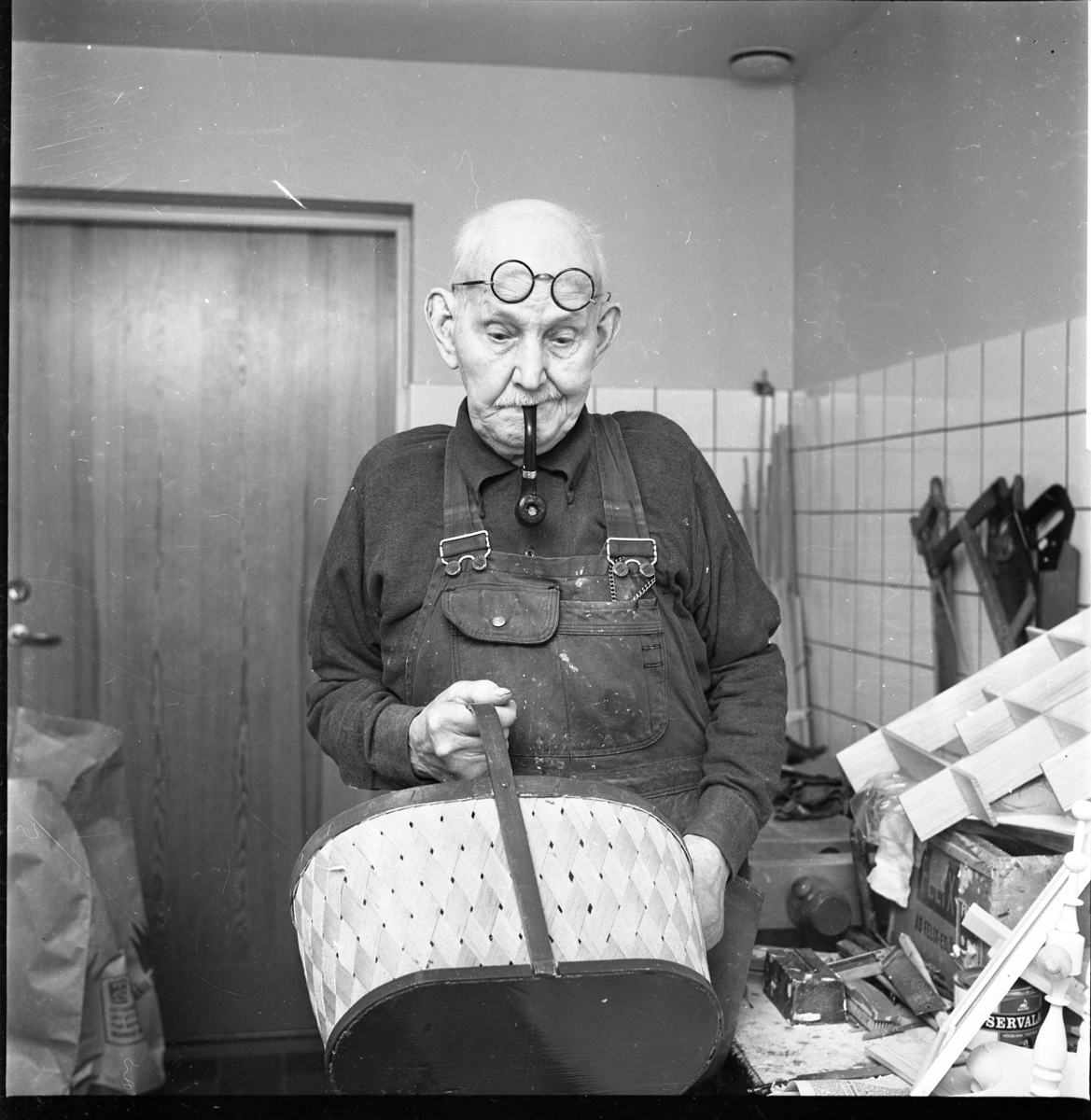Korgmakaren Emil Lund, 91 år, står i snickarbyxor med runda glasögon uppdragna i pannan och pipa i munnen. Han håller en korg i en handen och tittar ned mot den.