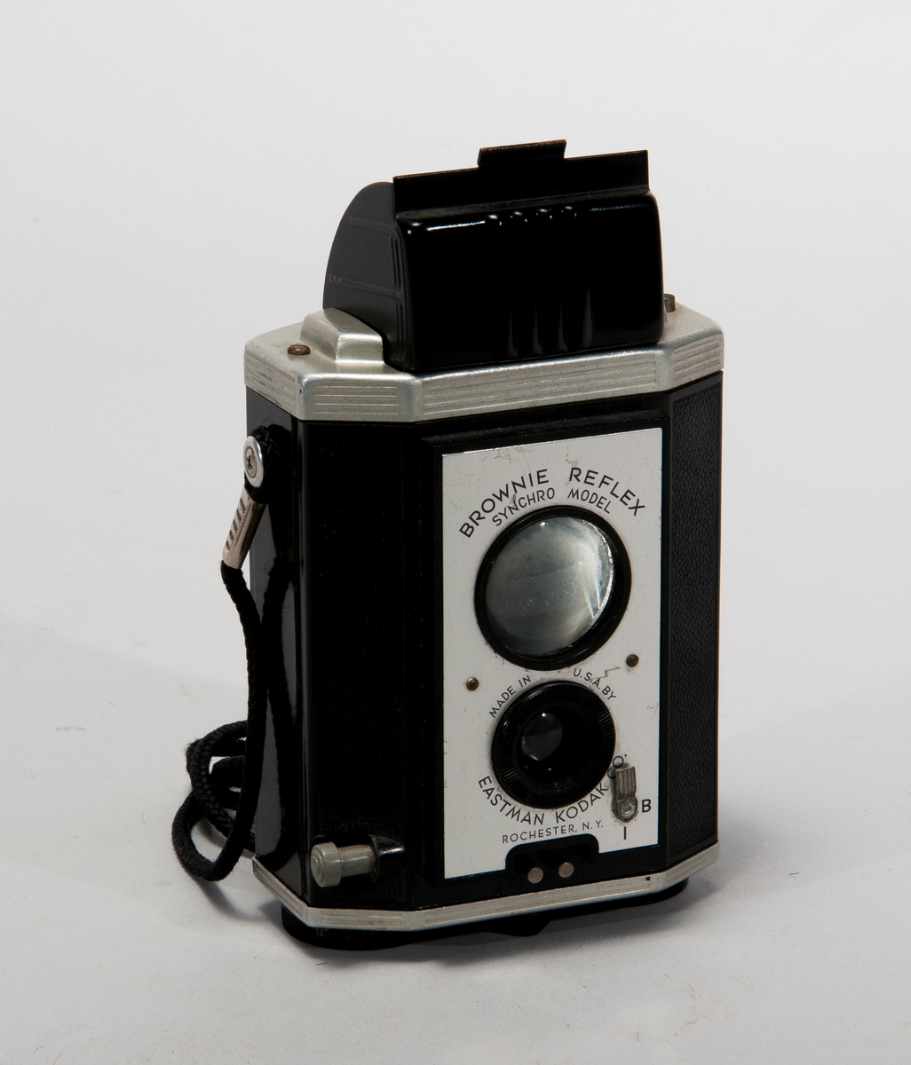 Tvåögd spegelreflexkamera för mellanformat, tillverkad i bakelit och metall. Med axelrem i textilmaterial.
Märkt: Brownie Reflex Synchro Model.