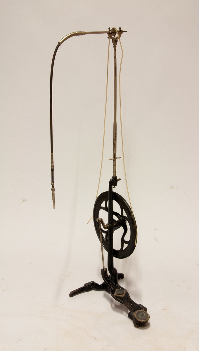 Svart tandläkarborr med svänghjul i gjutjärn. Drivs med en pedal. borren hänger i änden av ett böjligt rör. Märkt "1895 D.R.G.M" uppe vid röret.