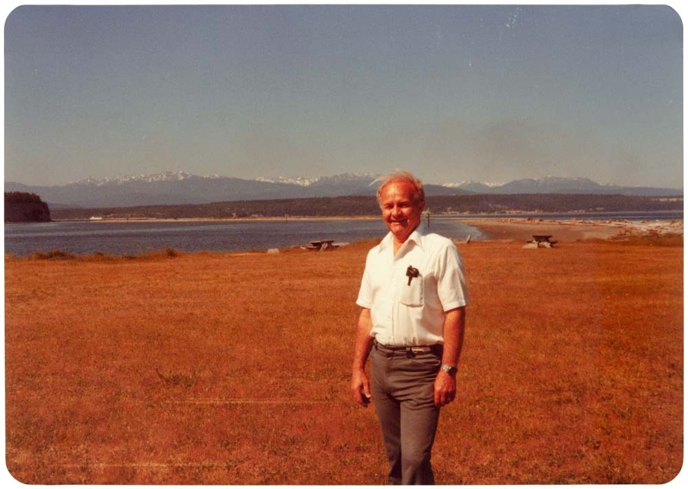 Mann står ved innsjø med fjell i bakgrunnen. "Dick 1979, Port Townsend across the water"