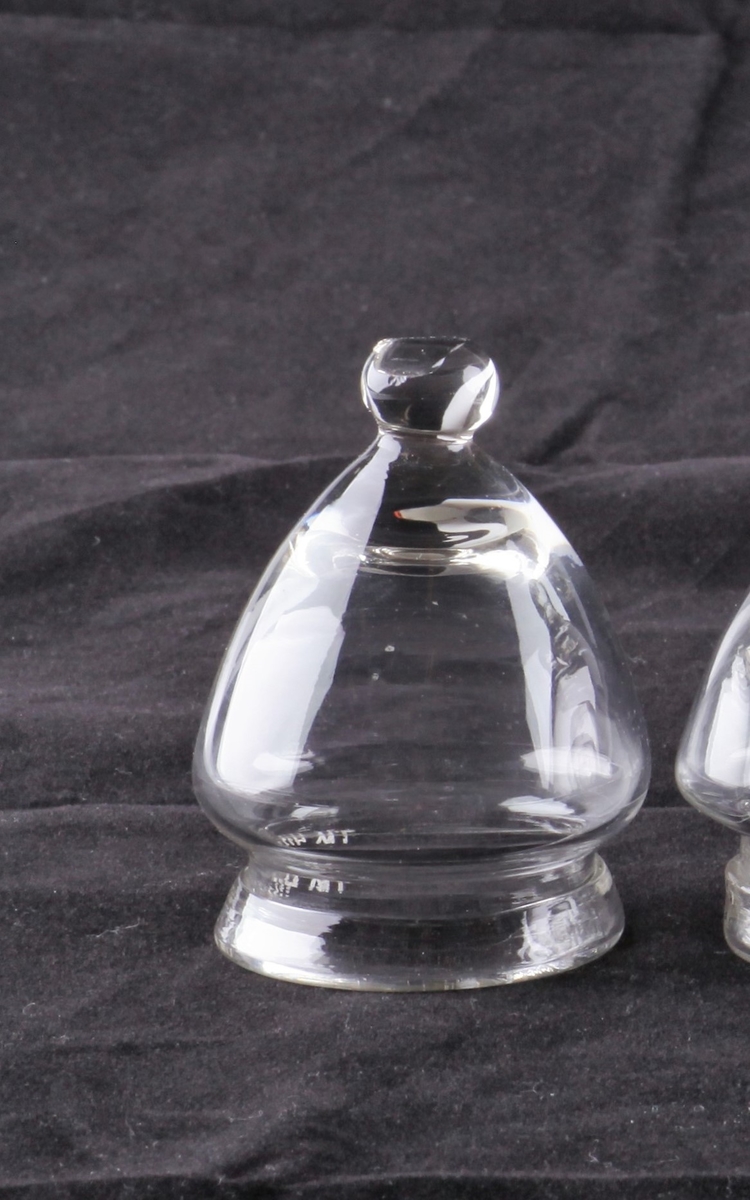 Koppeglass blåst i blankt glass. Formet som en klokke med en rund knapp øverst, og med glassmassen brettet ut som en brem nederst.
