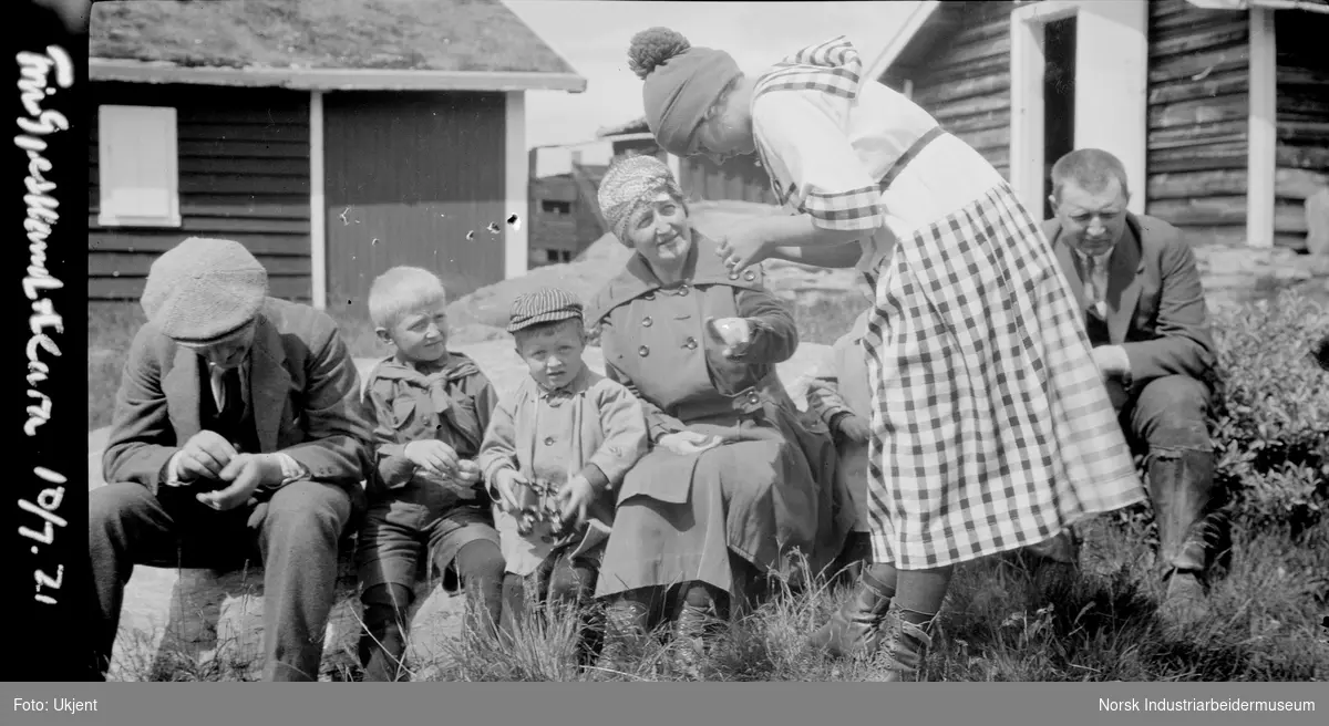Familie på tunet ved fjellbruk. To menn, en kvinne og tre barn sitter på rekke. En kvinne i rutete kjole står bøyd foran menneskene.
