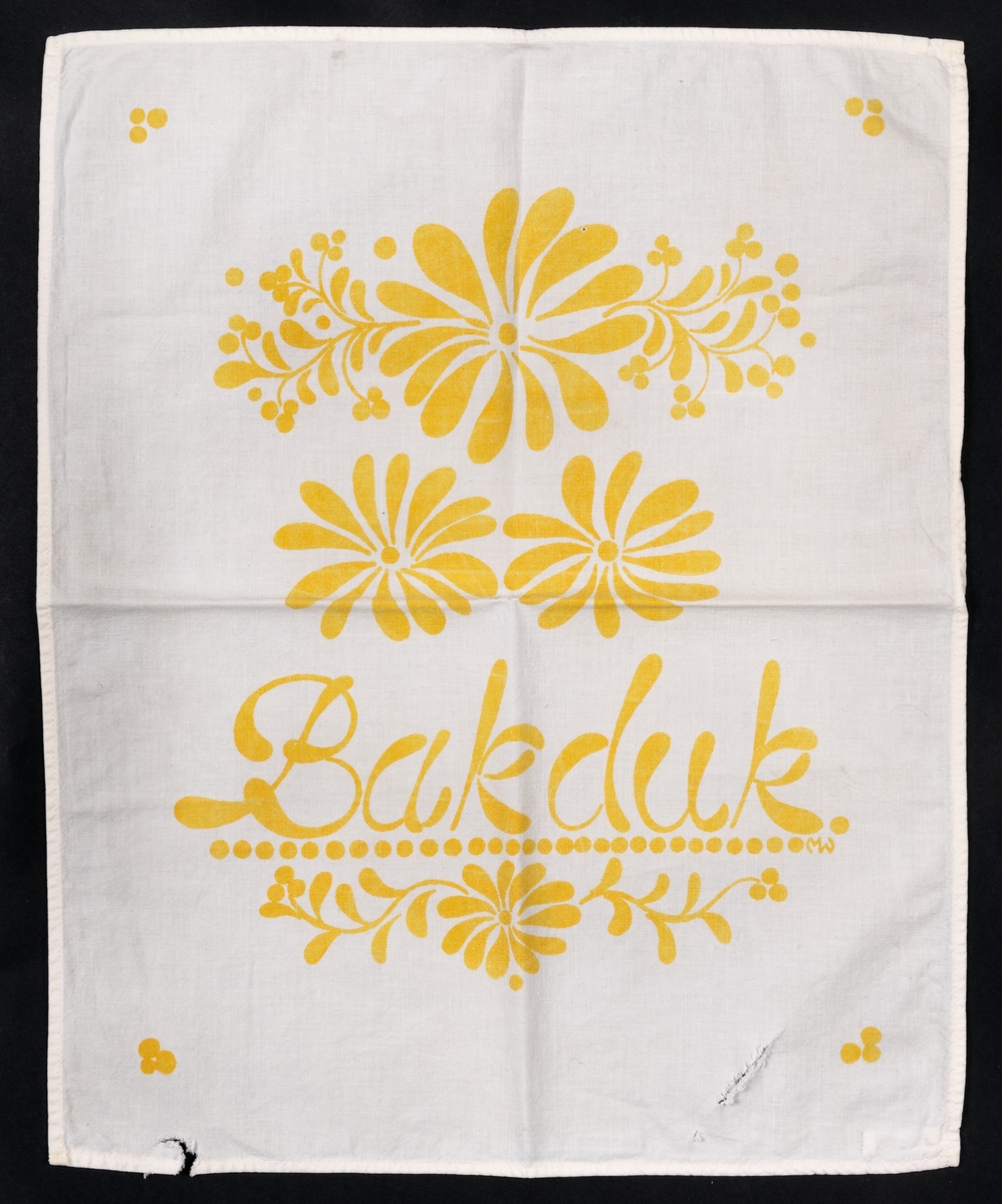 Bakdduk av vitt, maskinvävt bomullstyg med tryckt mönster i gult. Handduken är från 1970-talet men mönstret är inspirerat från sekelskiftet 1900. Hål i ena sidan.