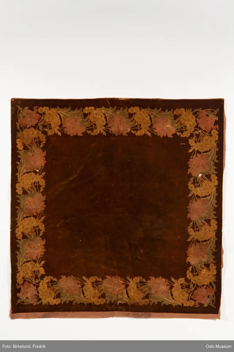 Gull- brun fløyelsduk fra slutten av 1800- tallet (ant.)
Duken er kvadratisk med blomstermotiv langs kantene, i lyse duse farger i grønn, gul og rosa. Baksiden er trukket med et grovt brunt stoff.