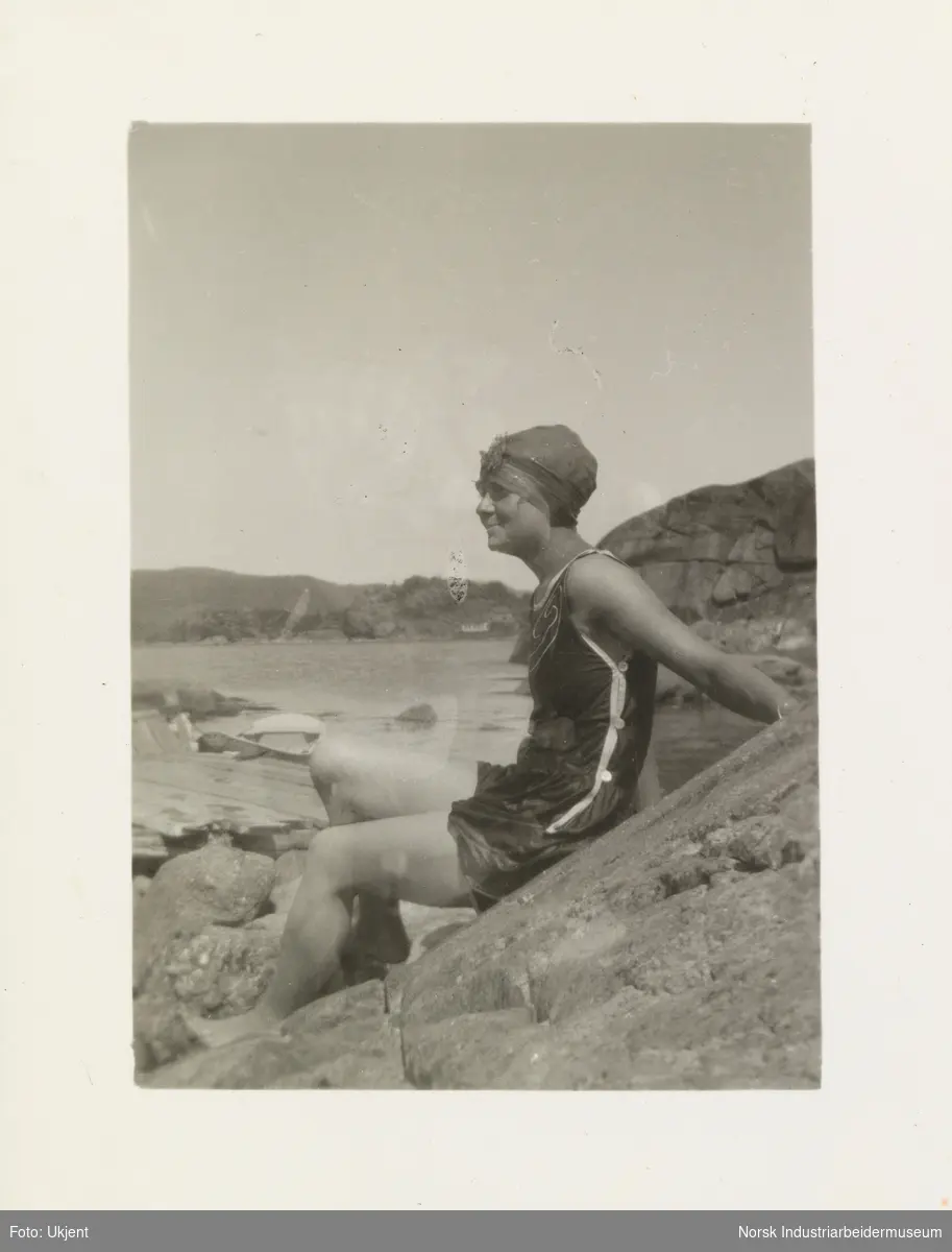 Kvinne i badrakt og lue sitter på svaberg etter et bad i sjøen
