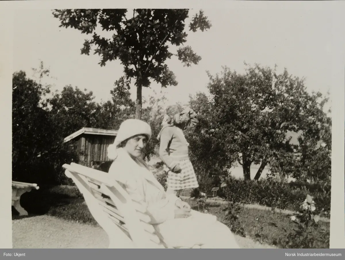Kvinne sitter utendørs på en hagebenk. Ved siden av står en jente med skaut på hode og speider