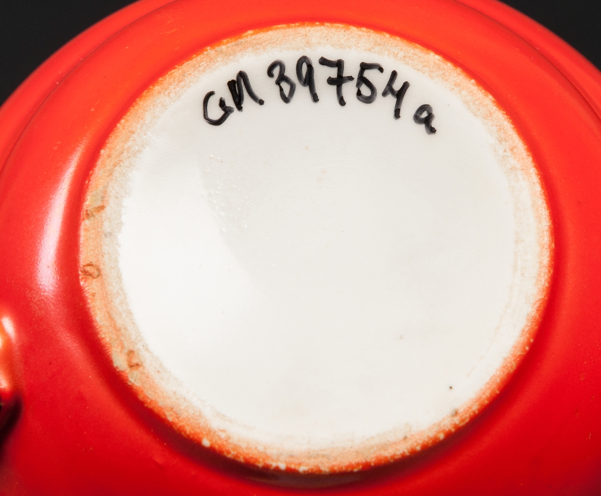 Kaffekopp med fat. Röd glasyr med svarta kanter och svart hänkel. Insidan av koppen vit, samt undersidan av fatet.