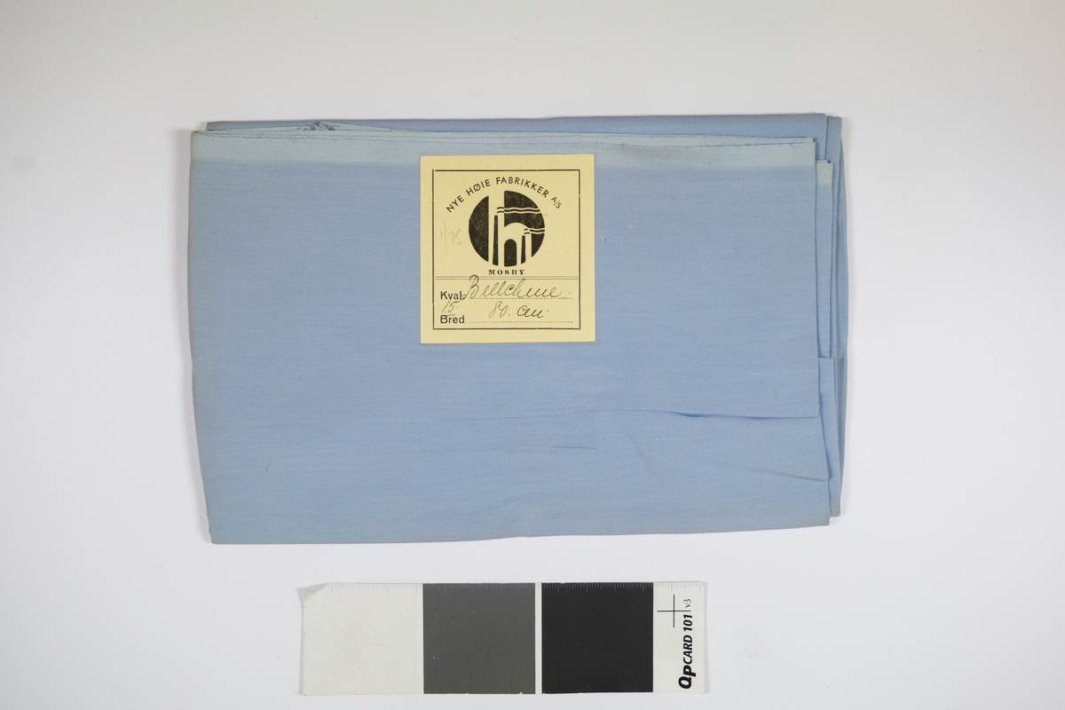 Tekstilprøve av sammenfoldet stoff, med påklistret papirlapp. Ensfarget tynt blått tekstil. Et papirkort ligger inni tekstilet.