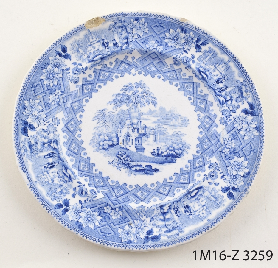Rund vit assiett med tryckt blå dekor föreställande (mittmotiv) landskap och ornamenterad bård på bräm och kepkant.