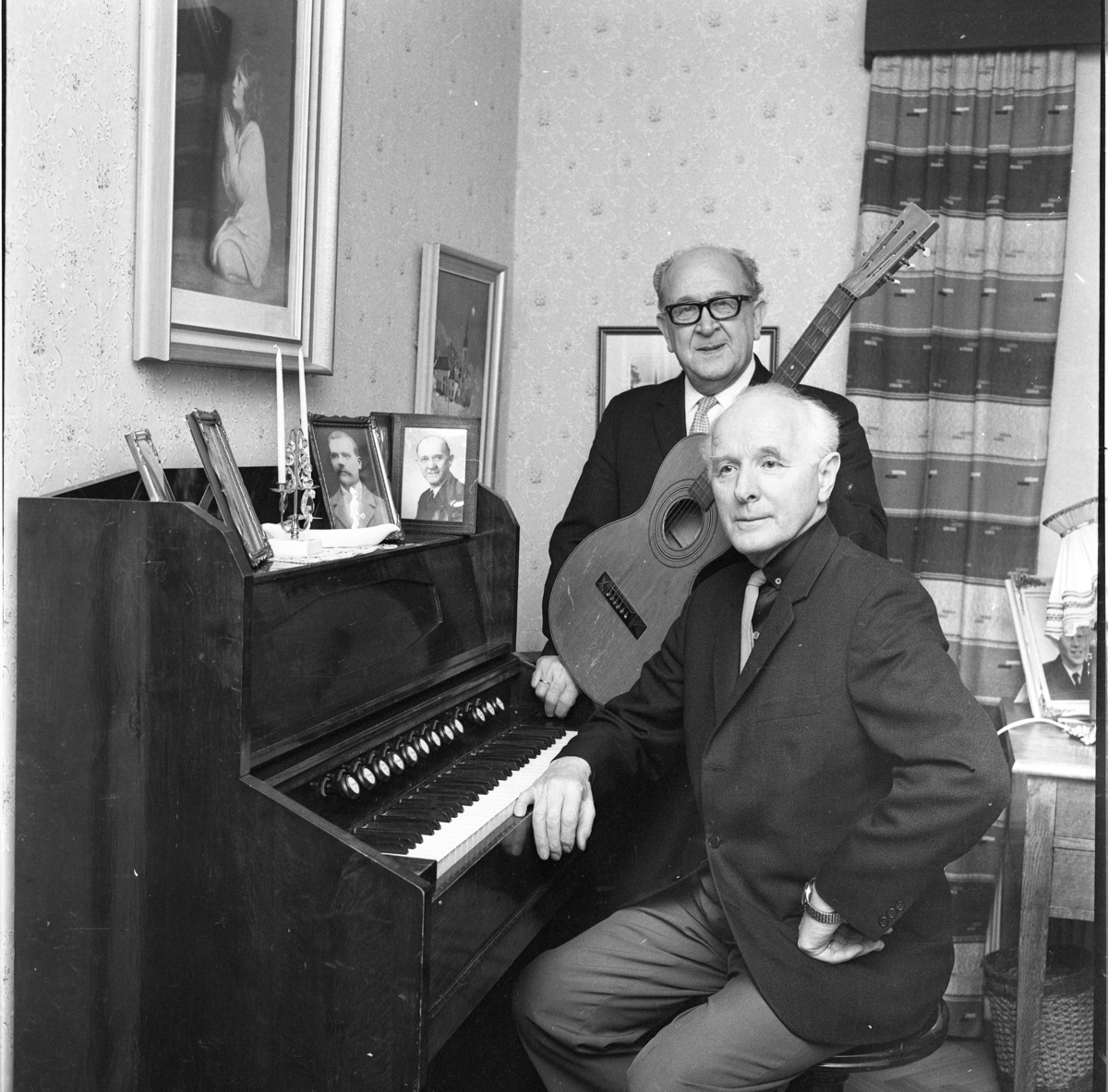 Carl Öst i glasögon står med en gitarr intill Martin Persson som sitter vid en orgel. På orgeln står en ljusstake med två ljus samt ramade porträttfotografier av män.