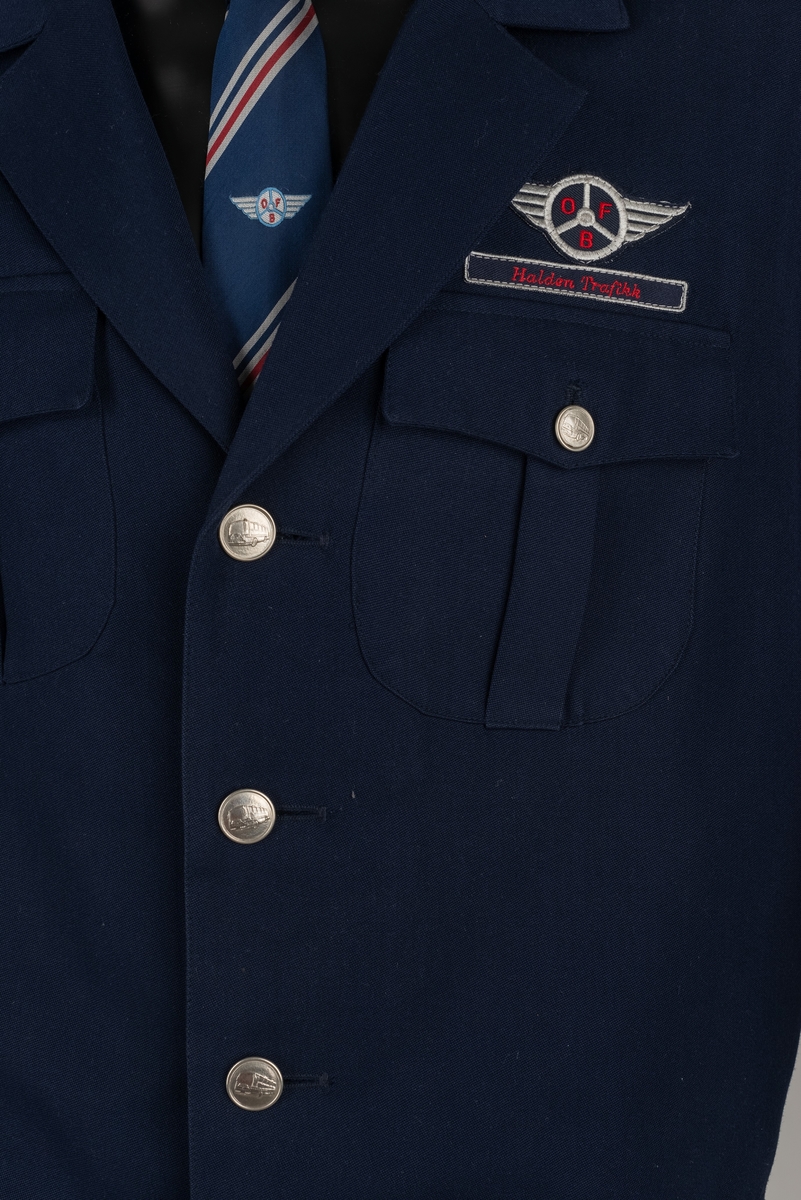 Uniformen består av bukse og jakke i mørk blå ull og polyester. Slips i kunstsilke, mørkeblått med hvite og røde diagonale striper. Pengeveske i ubehandlet lær.