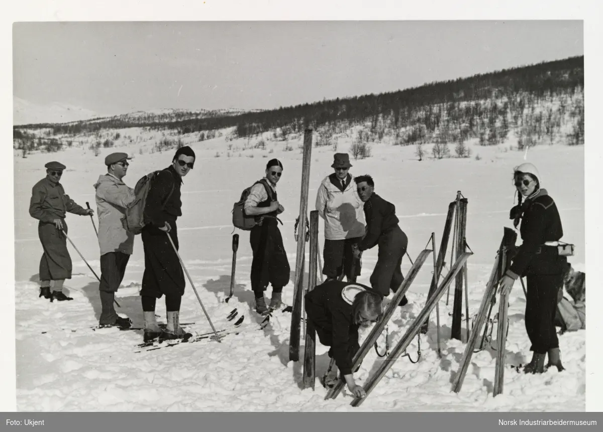 Gruppe med mennesker i turantrekk og solbriller gjør seg klar til skitur, påsken 1940. Noen av menneskene har på seg skiene, to jenter smører og gjør klar skiene.