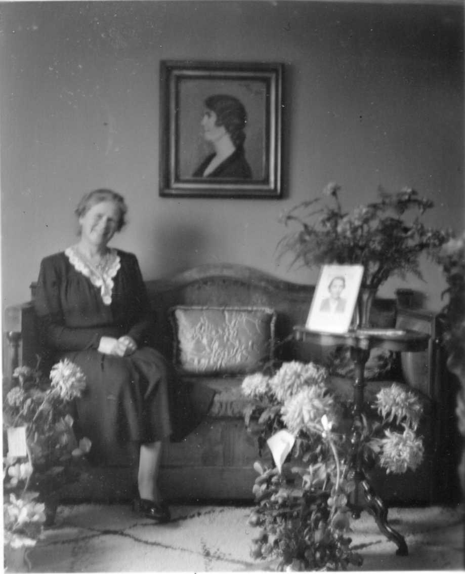 Anna Kellner i mörk klänning med spets vid halslinningen, sitter i en soffa med blomsterkorgar stående på en matta på golvet och i en vas på ett pelarbord. På bordet står också ett porträttfotografi i en ram. På väggen ovan soffa hänger ett kvinnoporträtt i profil. Det är sannolikt Anna Kellners födelsedag.