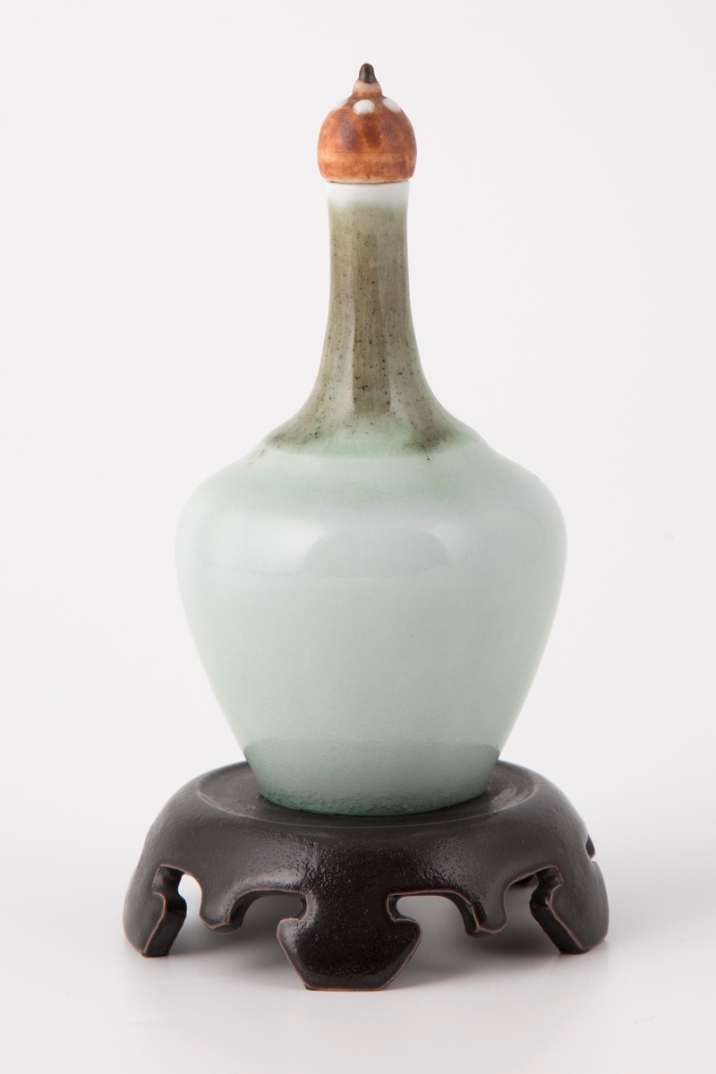 Porselensvase som hviler på en lav fot av steingods. Flaskeformet korpus med lang og smal hals. Korpus er dekket av hvitfarget glasur med et grønnlig skjær, mens halsen er grønn med spetter. På toppen av halsen sitter en kuleformet kork, hvis brunfargede overflate er besatt med fem små dråper av hvit glasur. Vasen er skrudd fast til en firebent fot, som er sortbrun med orientalsk-inspirert utforming.