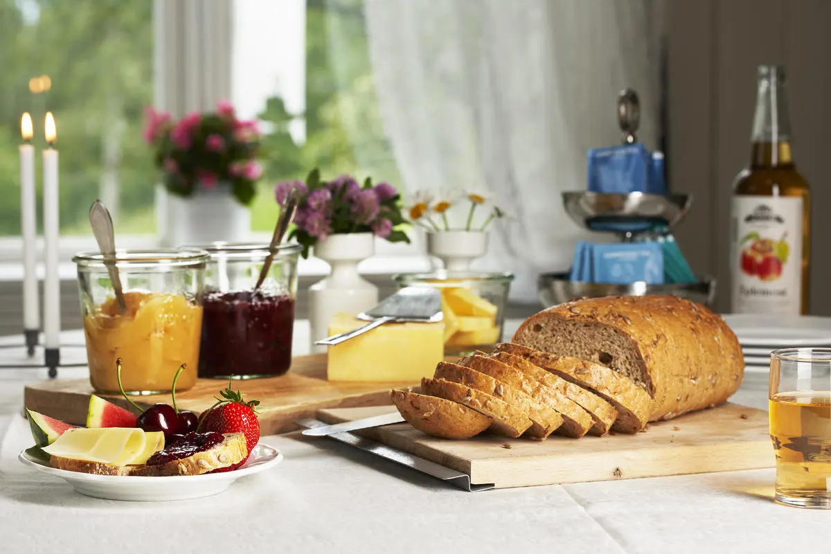 Et pent dekket frokostbord med brød, eplemost, syltetøy og ost. Blomster og levende lys i bakgrunnen