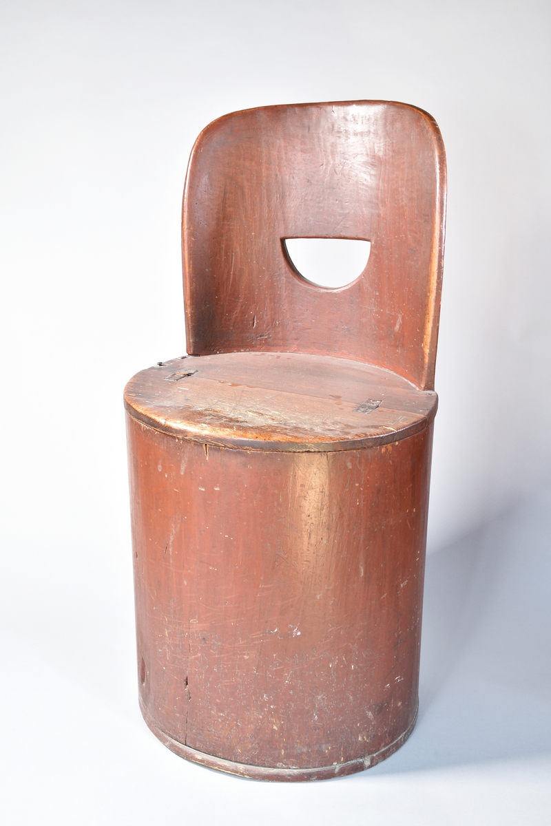 Kubbstol, urholkad av furu, målad rödbrun. Svängt ryggstöd med halvcirkelformat urtag för handgrepp. Halva sitsen uppvikbar med gångjärn av läder.