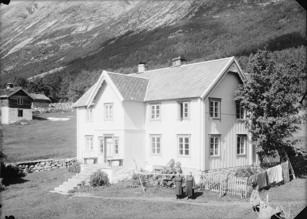 Stort velholdt hus med liten hage ved foten av bratt fjellside. To kvinner står ved huset. Glimt av lite tømmerhus på høy steinmur