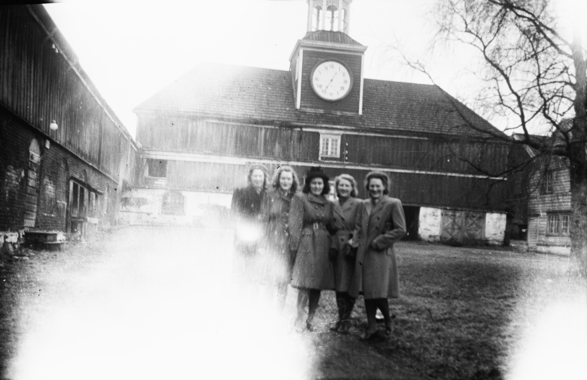 Gruppeportrett av fem kvinner som står i en gårdsplass, foran en bygning med et større klokkeur.