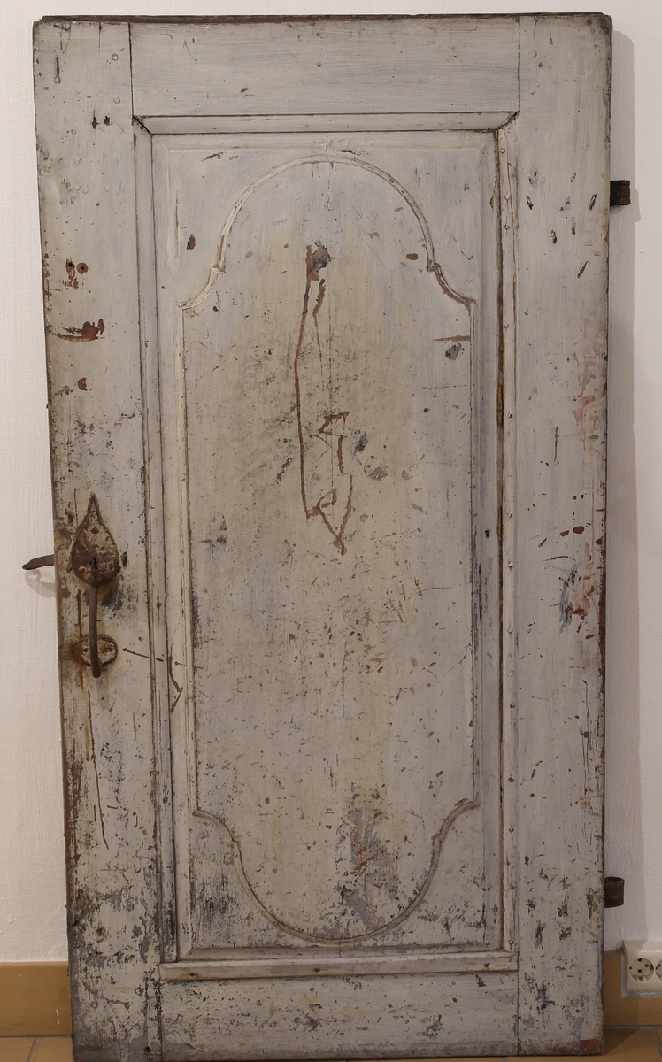 Dør i furu med smidde jernbeslag og hengsler. Døra er opprinnelig rødmalt, og malt grå over i ettertid. På dørspeilet er det malt en tekst.