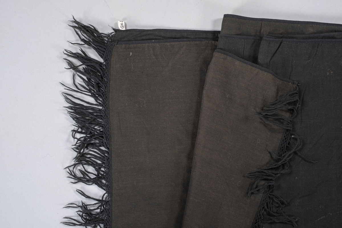Rektangulært sort sjal i bomull. På begge kortsidene er det en flettet kant med knyttede frynser ytterst. Sjalets underside er litt lysere enn oversiden.