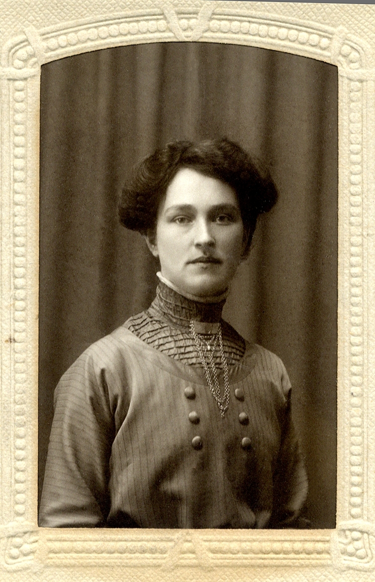 Foto av en kvinna i mörk, höghalsad blus, med ett halslås med kedjor vid kragen. 
Bröstbild, halvprofil. Ateljéfoto.
