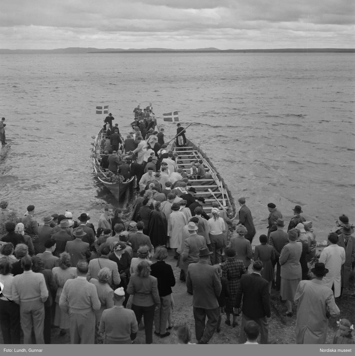 Motiv: (ingen anteckning) ;
En folksamling vid Rättviks kyrka, en folksamling står vid en brygga när kvinnor och män går ombord på två kyrkbåtar.