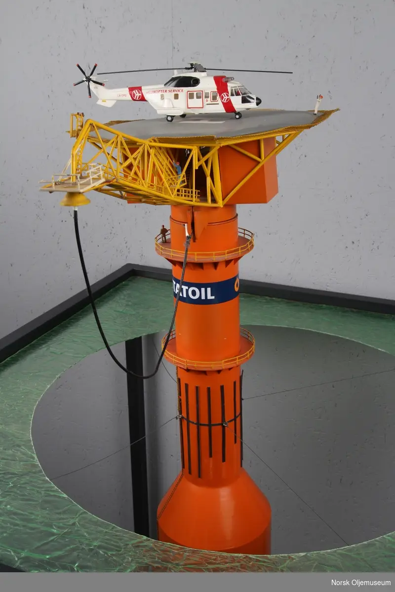 Modell av lastebøyen knyttet til Statfjord A. Malt orange og gul med helikopter på helidekket. Modellen holdes på plass av en aluminiumsramme der bunn og topp forestiller hhv. havbunn og hav.