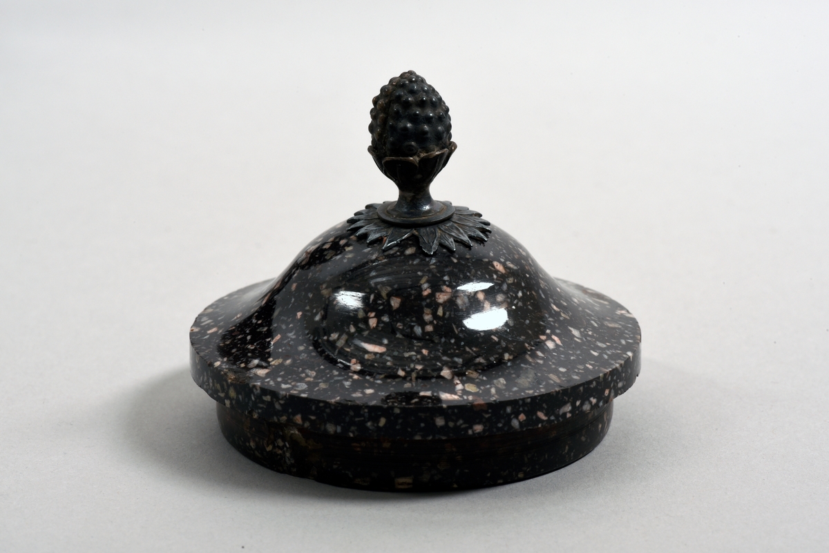 En av två urnor med lock av porfyr från Älvdalen. Kvadratisk fot. Locket har en knopp i form av en bladkransad pinjekotte av silver. Urnan är i tre isärtagbara delar.