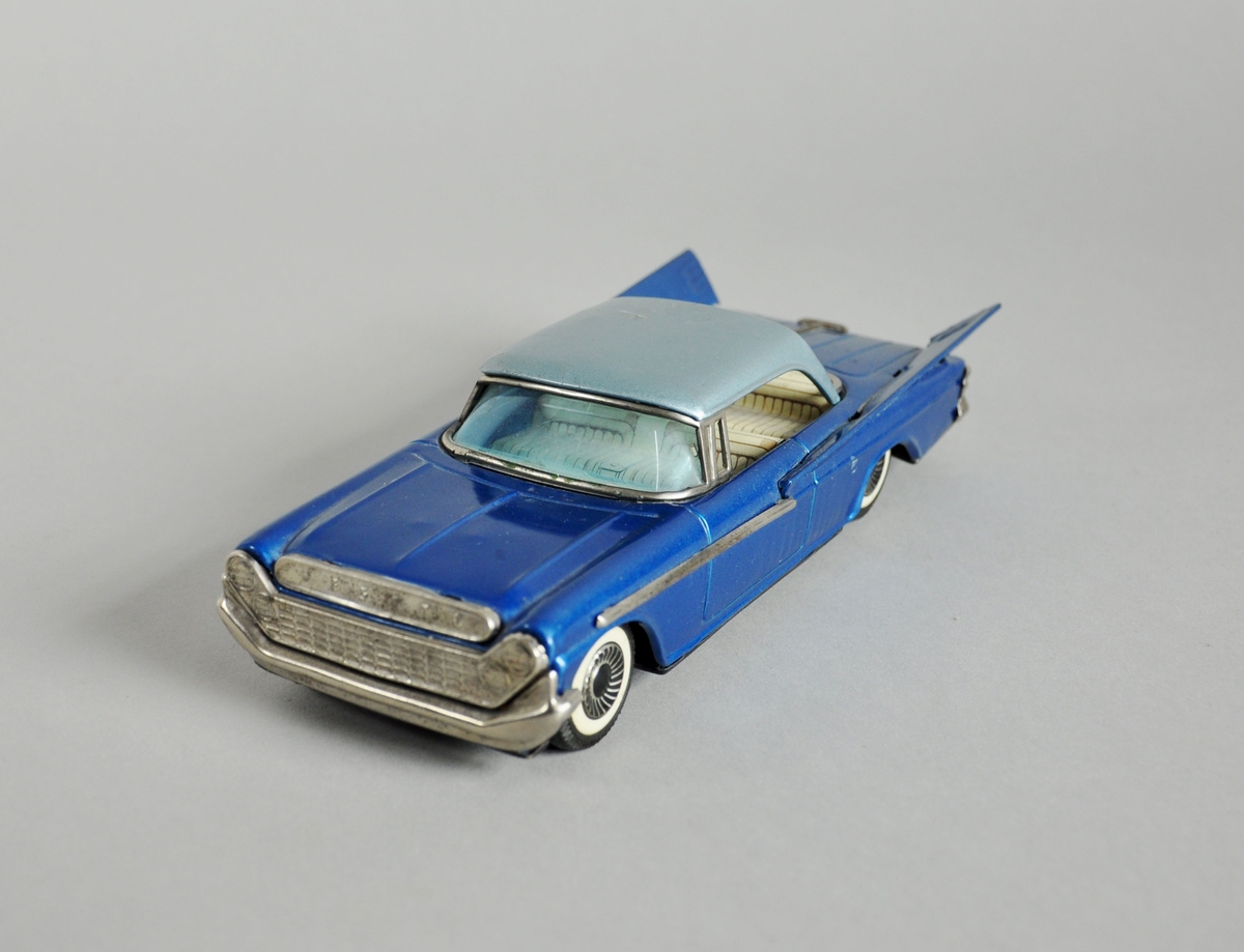 Blå lekebil - amerikansk 1950-tall - med vinger på hver side bak. Bilen har hvite seter.