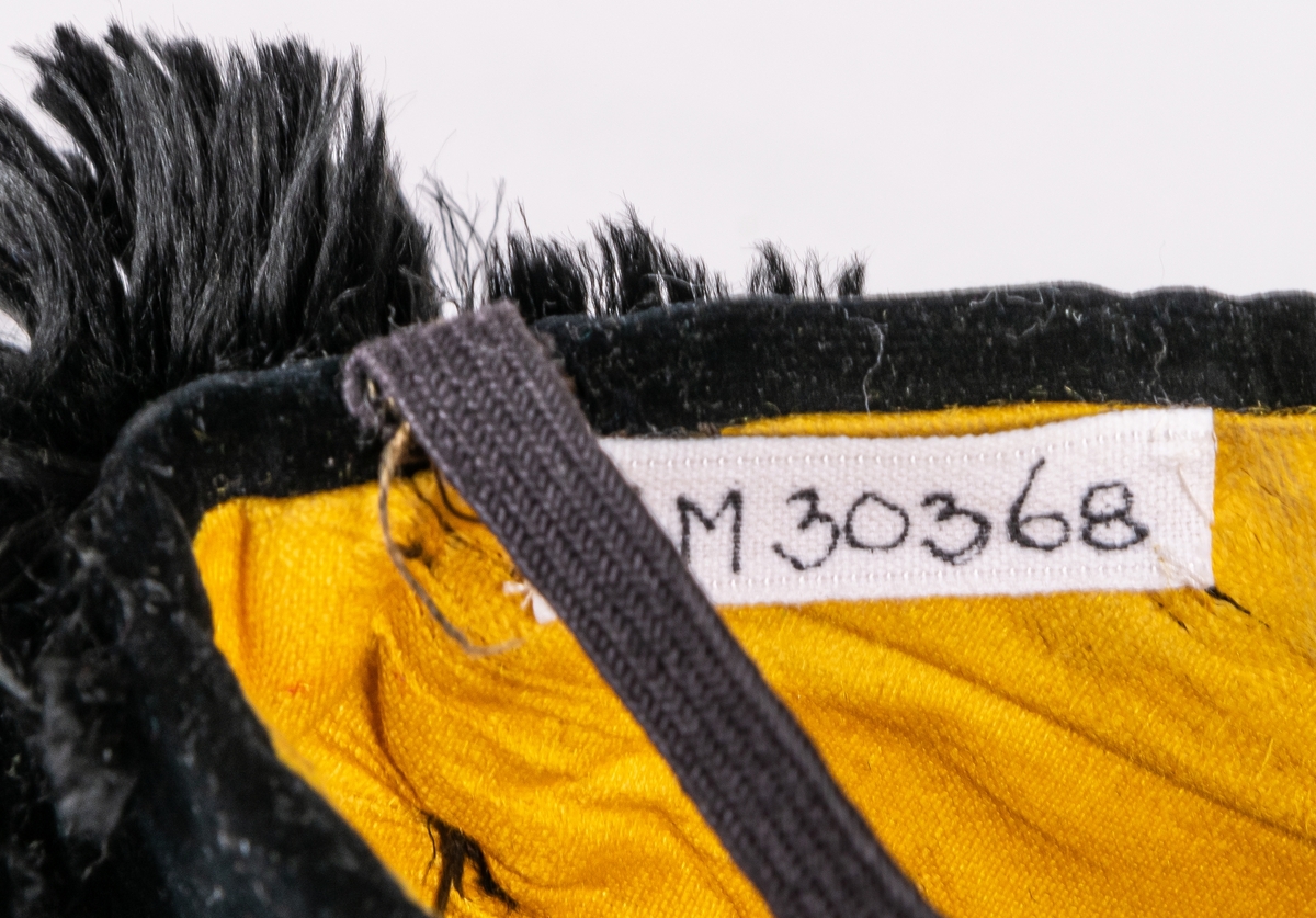 Mössa av svart sammet fodrad med gult bomullstyg. I sidorna svarta och gula fjädrar. Ett svart knytband. Hör till cirkusdräkten 30356.