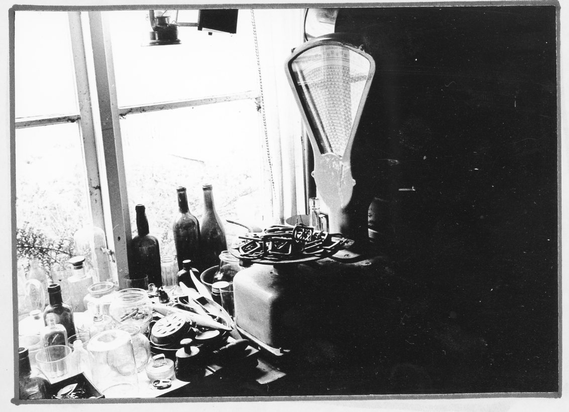 Bild från en lagerlokal på gården hos antikvitetshandlaren Carl Idh på Bergsgatan 60 i Gränna. En våg står på ett bord vid ett fönster med många andra prylar. På fönsterbrädan står glasflaskor.