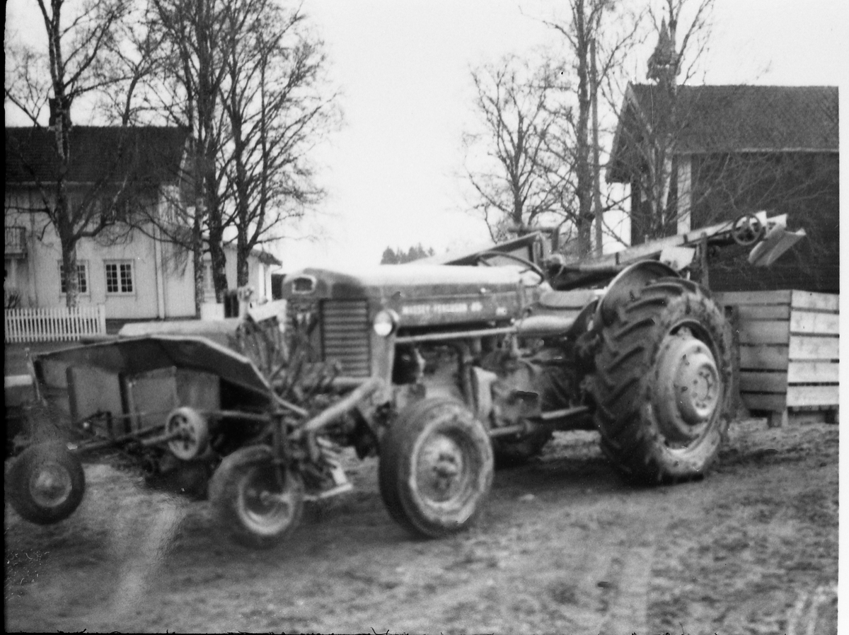 Avfotografert bilde av en traktor med potetopptaker som synes å være delvis frontmontert, men med transportbånd til potetkasse bak. Traktoren står på et gårdstun, og i bakgrunnen er et våningshus og et stabbur. Stedet er ikke identifisert.