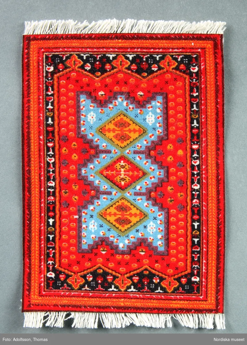 Matta i miniatyr föreställande en orientalisk matta. Mattan är röd, blå och svart med tryckt mönster. Den ska ligga vid den röda soffgruppen (NM.0333291 a-d), under det bruna pelarbordet i dockskåpets salong (NM.0333480).