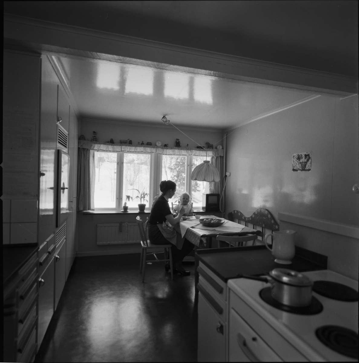villa Ahlgren
Interiör av kök. Kvinna med barn i motljus mot fönster i fonden