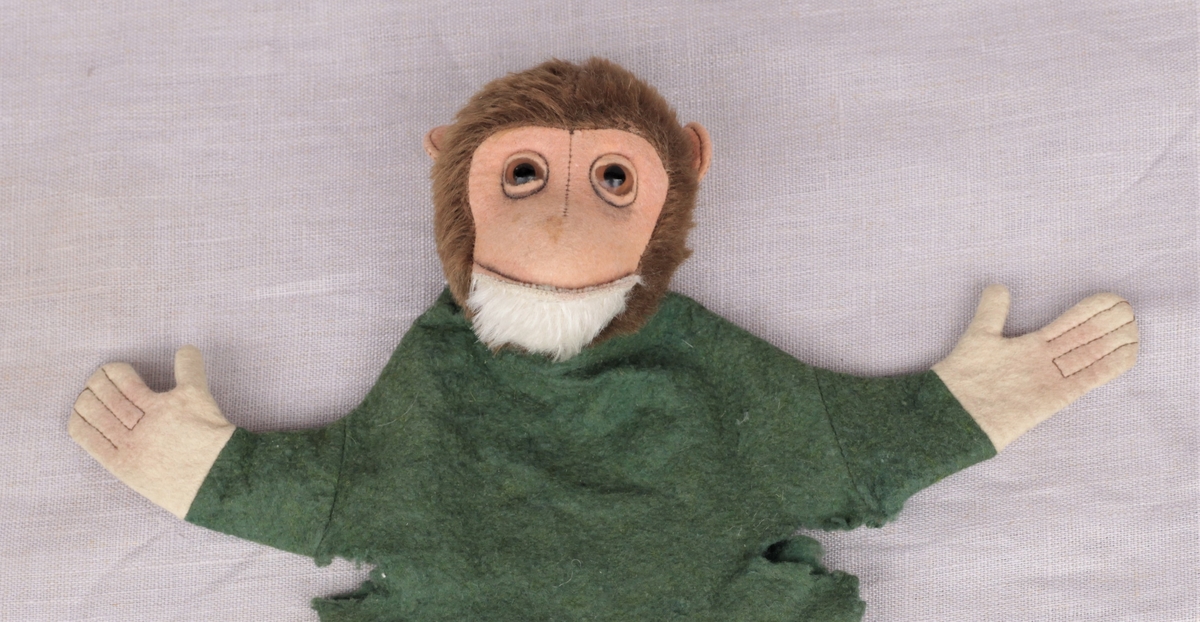 Hånddukke med fjeset til en apekatt, og grønn overdel. Apekatten har hvitt skjegg og brune øyne av glass.