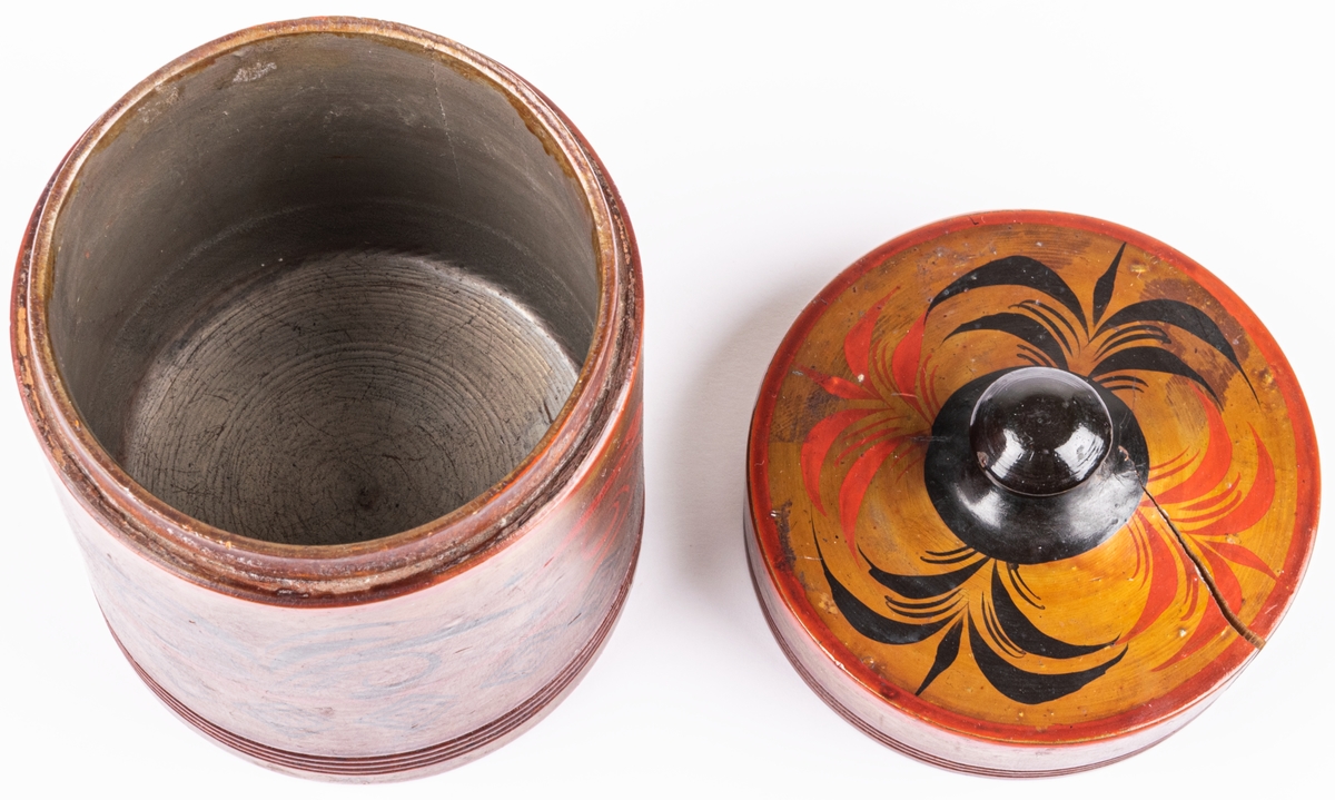 Kat.kort:
Tobaksburk av trä, svarvad, med knoppförsett lock, cylindrisk. Gulbrun bottenfärg med dekor i svart och rött.
