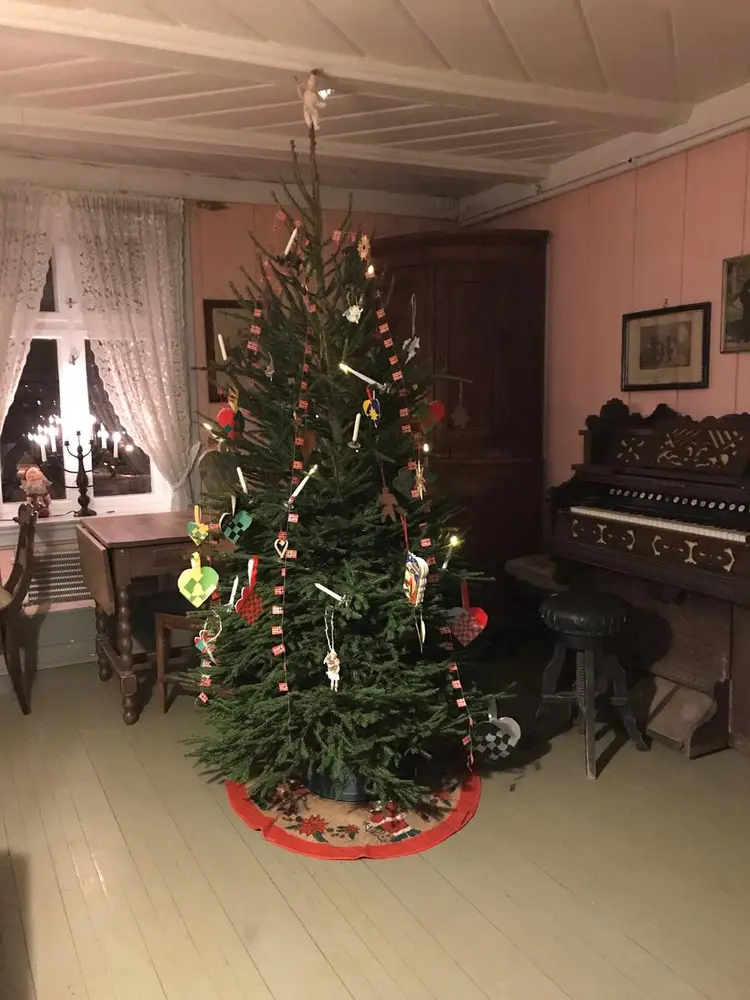 Juletre med gammeldags julepynt i finstua står foran et piano og et vindu.