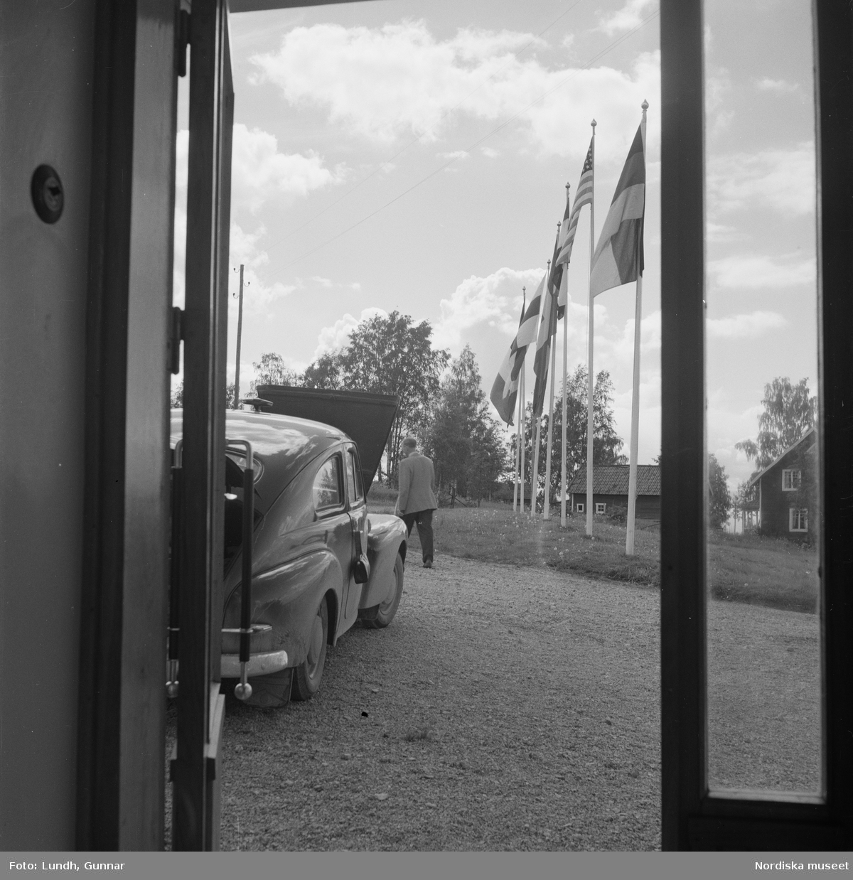 Motiv: Rättvik ;
Exteriör av hus, interiör med utsikt genom fönster med flaggstänger med nationsflaggor, tre kvinnor sitter vid ett trädgårdsbord vid parkerade bilar, porträtt av en man som sitter på en förstukvist, en kvinna och tre män spelar boccia.