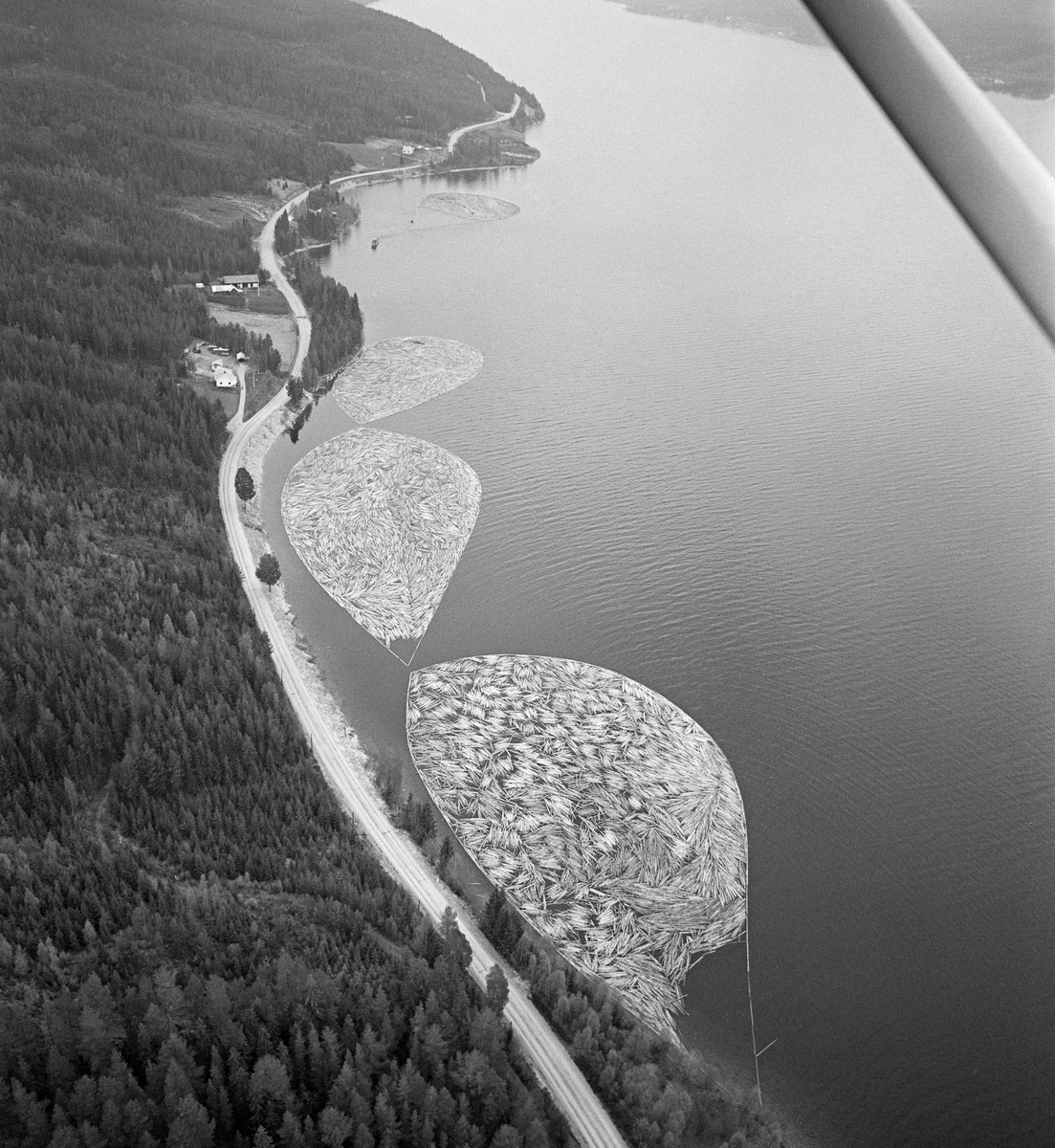 Fortøyde tømmerbommer ved land i Valmen, ei vik i den nordvestre enden av Osensjøen, der sjøen har avløp til elva Søndre Osa. Nordre Osen, Åmot kommune, Hedmark. Fotografiet er tatt fra fly.