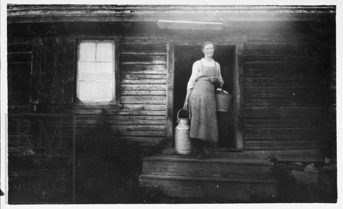Ei budeie står utenfor seterstørhus på Kollen seter i Helgedalen. Hun står med et mjølkespann og ei sinkbøtte.