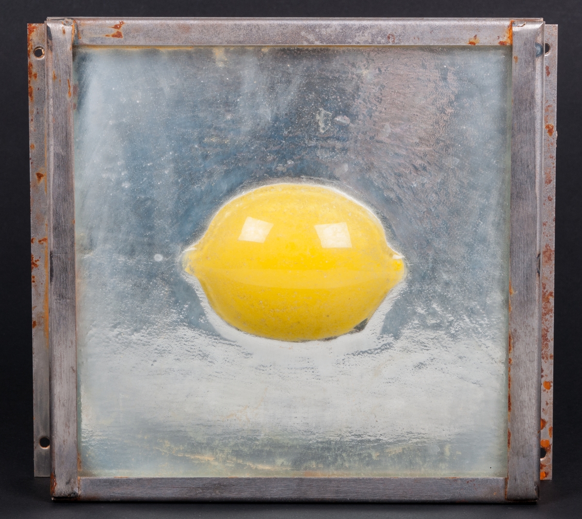 Objekt, skulptur, i glas och stål, "Spegelbild" (alt. Gul citron) av Lena Forslund-van Leer. Kvadratisk, något strukturerad, klar glasskiva med gul citron i relief i centrum. Bakom glasskivan är ett spegelglas. Glasskivorna infattade i obehandlad stålram.