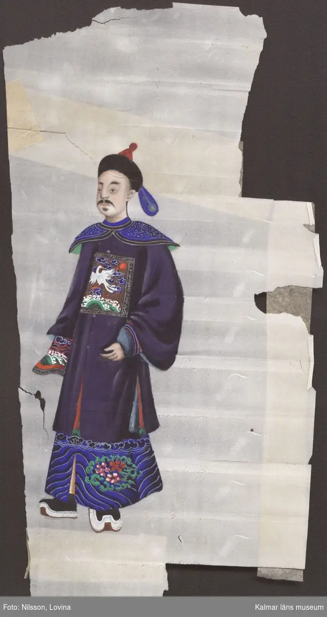 KLM 18866. Pappersmålning. Tunnt papper. Kinesisk pappersmålning i färg föreställande stående asiatisk man. Mannen bär en blå klädsel med olika mönsterutsmyckningar. På huvudet bär han en huvudbonad med en blå fjäder.